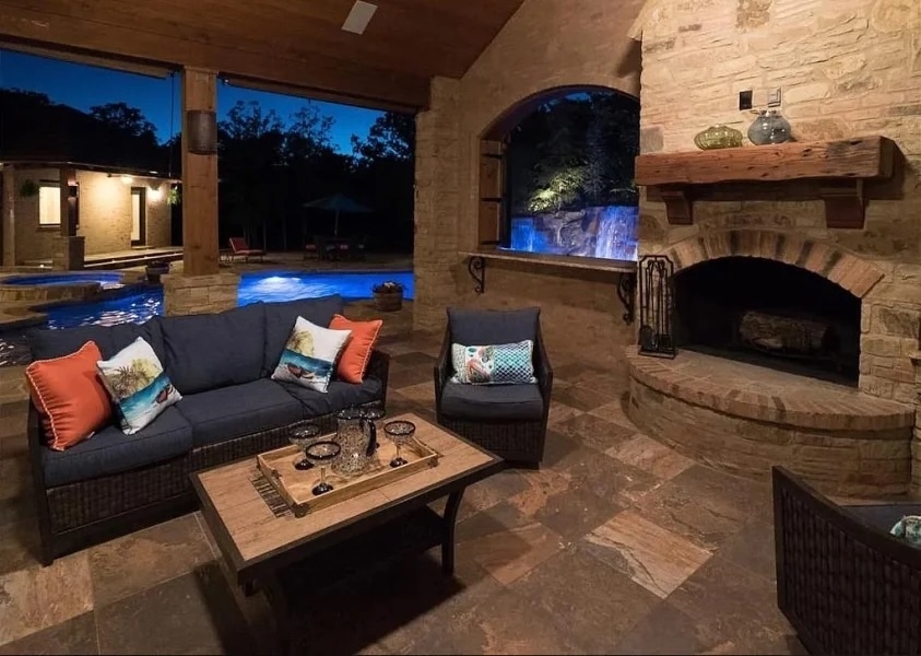 Elegant evening patio with a stone fireplace, outdoor seating, and a view of a glowing blue pool and waterfall.