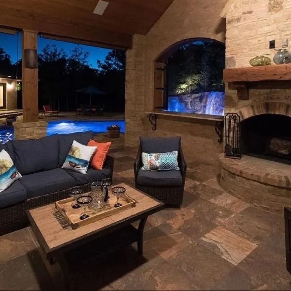 Elegant evening patio with a stone fireplace, outdoor seating, and a view of a glowing blue pool and waterfall.
