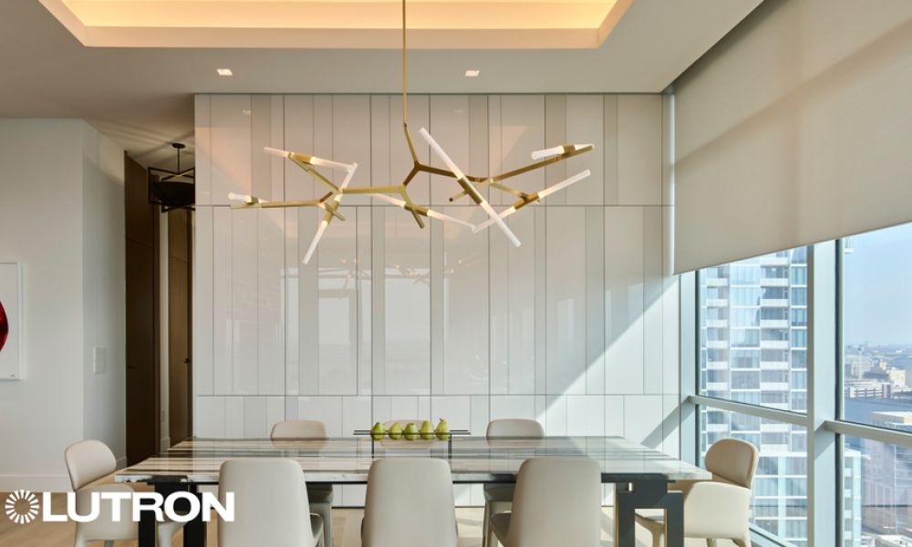Modern dining room with a sculptural chandelier, glass dining table, upholstered chairs, and large windows with automated window shades, featuring Lutron lighting control branding.