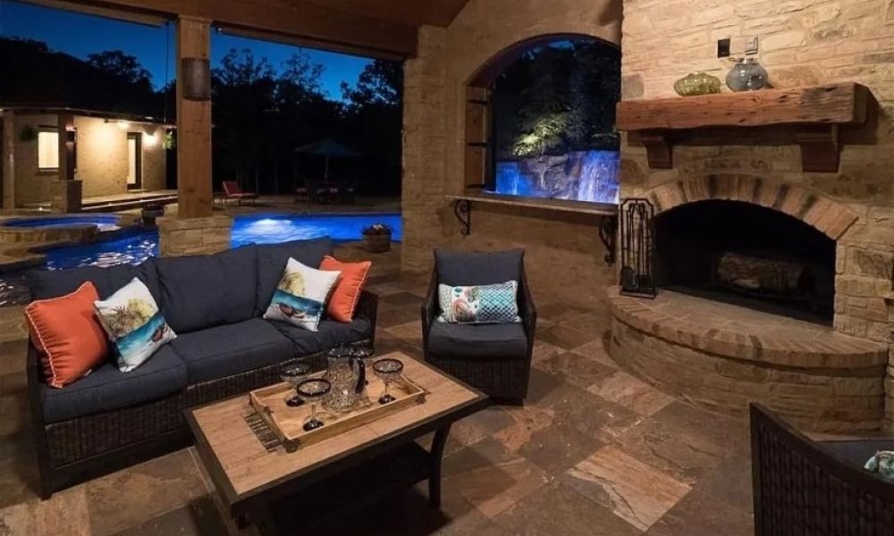 Elegant evening patio with a stone fireplace, outdoor seating, and a view of a glowing blue pool and waterfall.